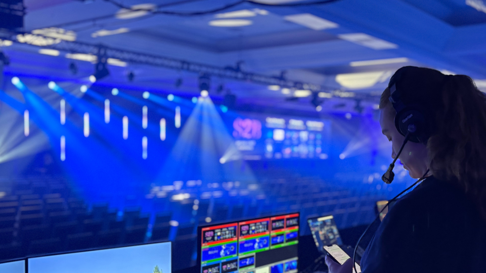 International event production in action: a producer with a headset monitors screens at the control desk of a conference hall with blue stage lighting.